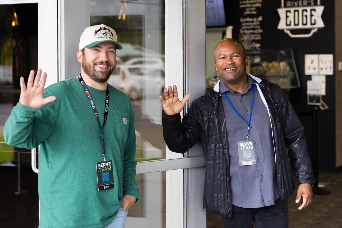Two smiling greeters wave hello at the front doors of The River